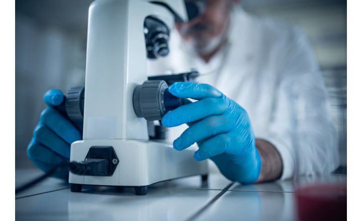 Microbiologist wearing white lab coat and blue nitrile gloves examples a sample using a microscope